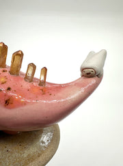 Ceramic sculpture of a pink dinosaur, mid stride, with orange quartz for spikes with a small character clinging to its tail.