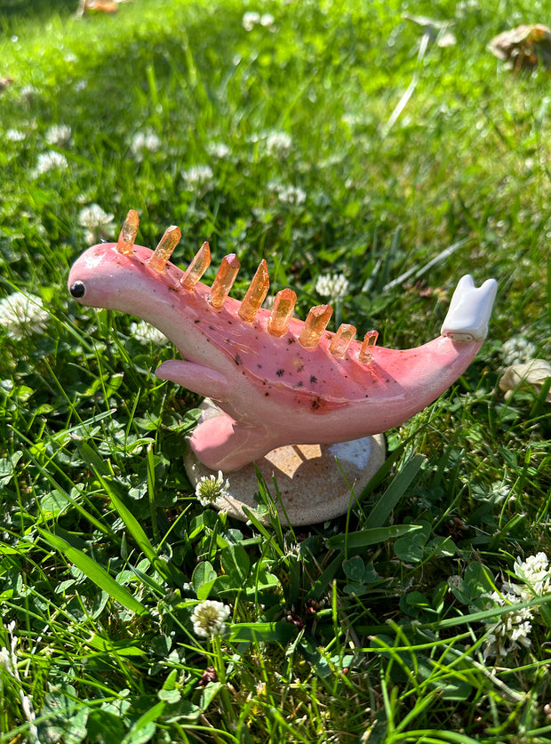 Ceramic sculpture of a pink dinosaur, mid stride, with orange quartz for spikes with a small character clinging to its tail.