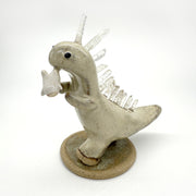 Ceramic sculpture of a cream colored dinosaur with large quartz on its back like spikes. It dangles a small cute character from its mouth by their foot.