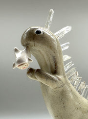 Ceramic sculpture of a cream colored dinosaur with large quartz on its back like spikes. It dangles a small cute character from its mouth by their foot.
