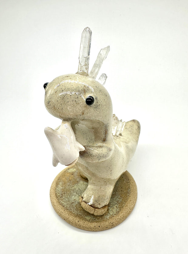 Ceramic sculpture of a cream colored dinosaur with large quartz on its back like spikes. It dangles a small cute character from its mouth by their foot.
