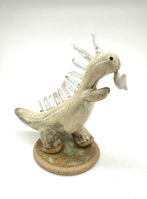 Ceramic sculpture of a cream colored dinosaur with large quartz on its back like spikes. It dangles a small cute character from its mouth by their foot.