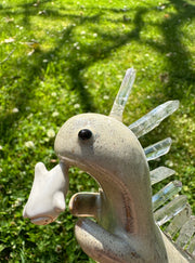 Ceramic sculpture of a cream colored dinosaur with large quartz on its back like spikes. It dangles a small cute character from its mouth by their foot.