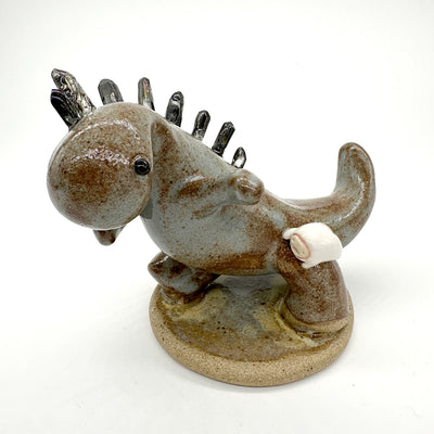 Ceramic sculpture of a brown and grey colored dinosaur with large quartz on its back like spikes. A small cute character clings on to the leg of the dinosaur.