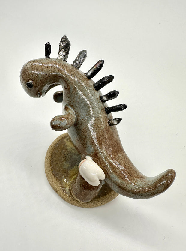 Ceramic sculpture of a brown and grey colored dinosaur with large quartz on its back like spikes. A small cute character clings on to the leg of the dinosaur.