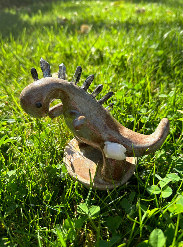 Ceramic sculpture of a brown and grey colored dinosaur with large quartz on its back like spikes. A small cute character clings on to the leg of the dinosaur.