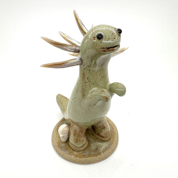 Ceramic sculpture of a brownish green colored dinosaur with curved spikes coming out of its neck like a collar. A small cute character clings on to the leg of the dinosaur.