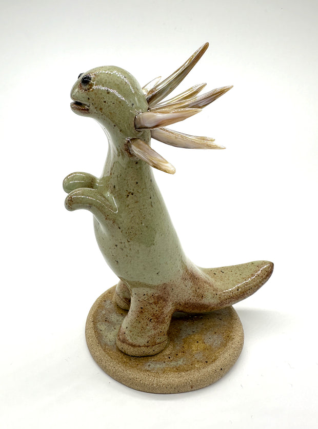 Ceramic sculpture of a brownish green colored dinosaur with curved spikes coming out of its neck like a collar. A small cute character clings on to the leg of the dinosaur.