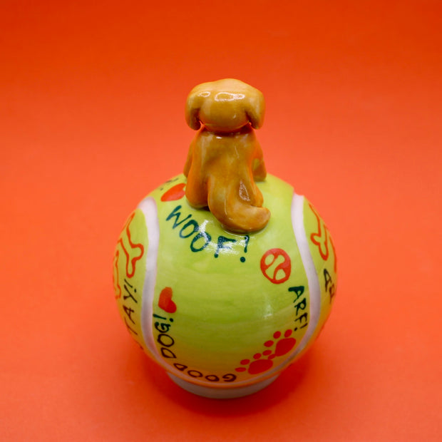 Ceramic sculpture of a small brown dog smiling and sitting atop a large tennis ball, with affirmations written on it with cute dog themed doodles.