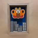 Shadowbox style artwork of a moth kaiju, floating over a cityscape with a blue background and light shining through.