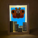 Shadowbox style artwork of a moth kaiju, floating over a cityscape with a blue background and light shining through.