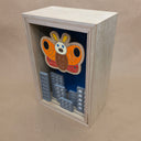 Shadowbox style artwork of a moth kaiju, floating over a cityscape with a blue background and light shining through.