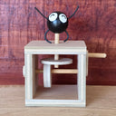 A wooden mechanical toy with a black round character figure on top, featuring white eyes and black thin arms and legs, set against a rustic reddish-brown wood background.