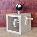 A wooden toy box with a crank handle and a black round character figure with white eyes and curved black arms and legs, set against a reddish-brown wood-grain background.