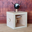 A wooden mechanical toy with a black round character figure on top, featuring white eyes and black thin arms and legs, set against a rustic reddish-brown wood background.