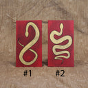 2 red envelopes made out of wood, with painted illustrations on each of gold snakes with black outlines. One snake is curved into itself, akin to the shape of an 8. The other is curved to make a multi-part "S" shape.
