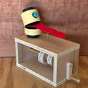 A wooden sculpture sitting atop a wooden box with a crank handle, featuring a yellow segmented head with a blue eye and a red tongue-like strip resting on metal pins, set against a wood-grain background.
