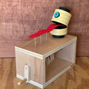 A wooden sculpture sitting atop a wooden box with a crank handle, featuring a yellow segmented head with a blue eye and a red tongue-like strip resting on metal pins, set against a wood-grain background.
