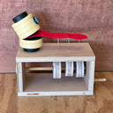 A wooden sculpture sitting atop a wooden box with a crank handle, featuring a yellow segmented head with a blue eye and a red tongue-like strip resting on metal pins, set against a wood-grain background.