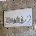 Laser wood cuts on a piece of ash wood of a series of buildings and a Godzilla character.