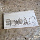 Laser wood cuts on a piece of ash wood of a series of buildings and a Godzilla character.