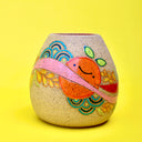 Ceramic sculpture on brown specked clay with a bright painting of an orange with a pink stripe running through it. Plants and other patterns are drawn as well.