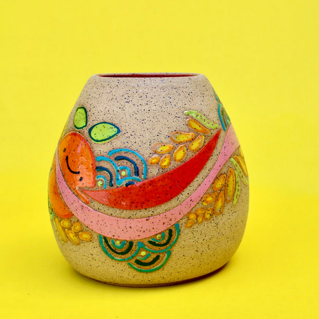 Ceramic sculpture on brown specked clay with a bright painting of an orange with a pink stripe running through it. Plants and other patterns are drawn as well.