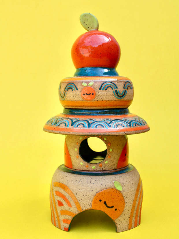 Sculpture made of 3 individual ceramic parts, stacked like a tower of sorts. The top of the tower is a bright round orange, atop of several small bowls and plate shapes. The bottom of the stack has a domed opening.