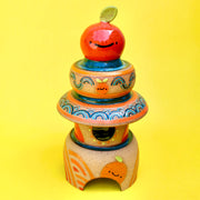 Sculpture made of 3 individual ceramic parts, stacked like a tower of sorts. The top of the tower is a bright round orange, atop of several small bowls and plate shapes. The bottom of the stack has a domed opening.
