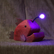 Whittled red wooden sculpture of an angler fish, with large cartoon type proportions. It has a light coming out of its head, emitting a purplish glow.