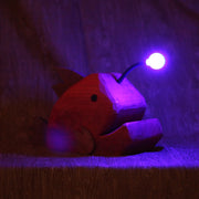 Whittled red wooden sculpture of an angler fish, with large cartoon type proportions. It has a light coming out of its head, emitting a purplish glow.