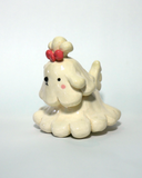 A small cream-colored ceramic figurine of a cute poodle or dog character with floppy ears, a pink bow on its head, and pink cheeks, set against a plain light background.