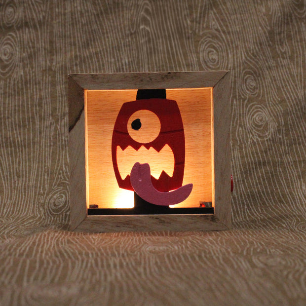 Wooden night light sculpture with a cut-out design of a face on a red lantern, with its tongue wildly out.