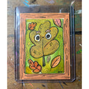 Artwork of a smiling leaf with cartoon eyes and stars on a green background, surrounded by autumn leaves. The artwork is enclosed in a clear protective sleeve and set against a rustic wooden background.