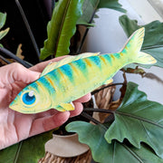 A hand holds a 6.75" long illustration of a green fish. It has striping down its body, large blue eyes, a wavy top fin, and a friendly, contented smile.