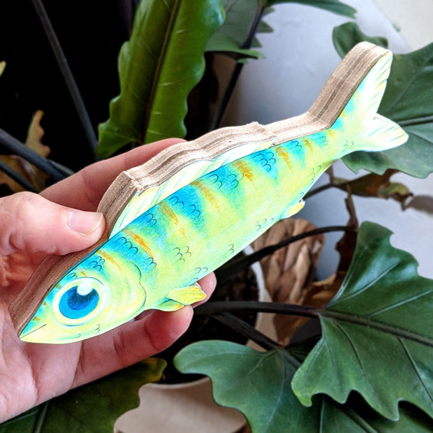 A hand holds a 6.75" long illustration of a green fish. It has striping down its body, large blue eyes, a wavy top fin, and a friendly, contented smile. At this angle we can see that the flat illustration is mounted on a thick piece of plywood, cut to the outline of the drawing.