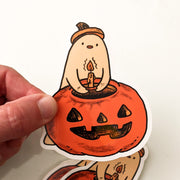 Die cut sticker of a cream colored ghost putting a candle into a jack o lantern.