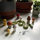 Collection of small sculptures, placed together to make a cohesive piece comprising of: a green snake and several different colored stout mushrooms.
