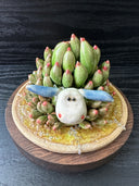 Sculpture of a white and blue sea slug with its back akin to a succulent, green with pink tips. It lays on a round ceramic base that has a green watery sheen.