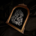 An ornate arched bronze framed mirror, featuring a silver-toned line art engraving of a woman's face surrounded by flowers, set against a dark draped fabric background.