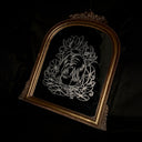An ornate arched bronze framed mirror, featuring a silver-toned line art engraving of a woman's face surrounded by flowers, set against a dark draped fabric background.