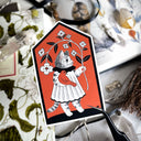 Sticker of a cat in a dress holding an orange bird and a large branch of flowers. Background is solid orange.