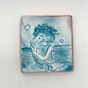 Blue and white illustration on a glazed terracotta tile of a girl standing in water, hugging a fish.
