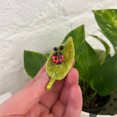 Small ceramic sculpture of a round ladybug on a green leaf. The lady bug has a cute smiling face.