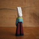Painted whittled wooden sculpture of a white rabbit with red eyes and a humanoid body, wearing a green and blue striped sweater and brown pants.