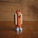 Small painted whittled wood sculpture of a dog dressed as a hot dog, with strips of mustard and ketchup. It wears a pair of silver shoes.