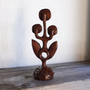 Whittled wooden sculpture of a statuesque plant, with only 2 leaves and 3 buds that have eyes on them. They all look off to the side and the plant is atop of a slightly spiked base. 