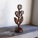 Whittled wooden sculpture of a statuesque plant, with only 2 leaves and 3 buds that have eyes on them. They all look off to the side and the plant is atop of a slightly spiked base. 