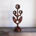 Whittled wooden sculpture of a statuesque plant, with only 2 leaves and 3 buds that have eyes on them. They all look off to the side and the plant is atop of a slightly spiked base. 