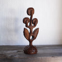 Whittled wooden sculpture of a statuesque plant, with only 2 leaves and 3 buds that have eyes on them. They all look off to the side and the plant is atop of a slightly spiked base. 